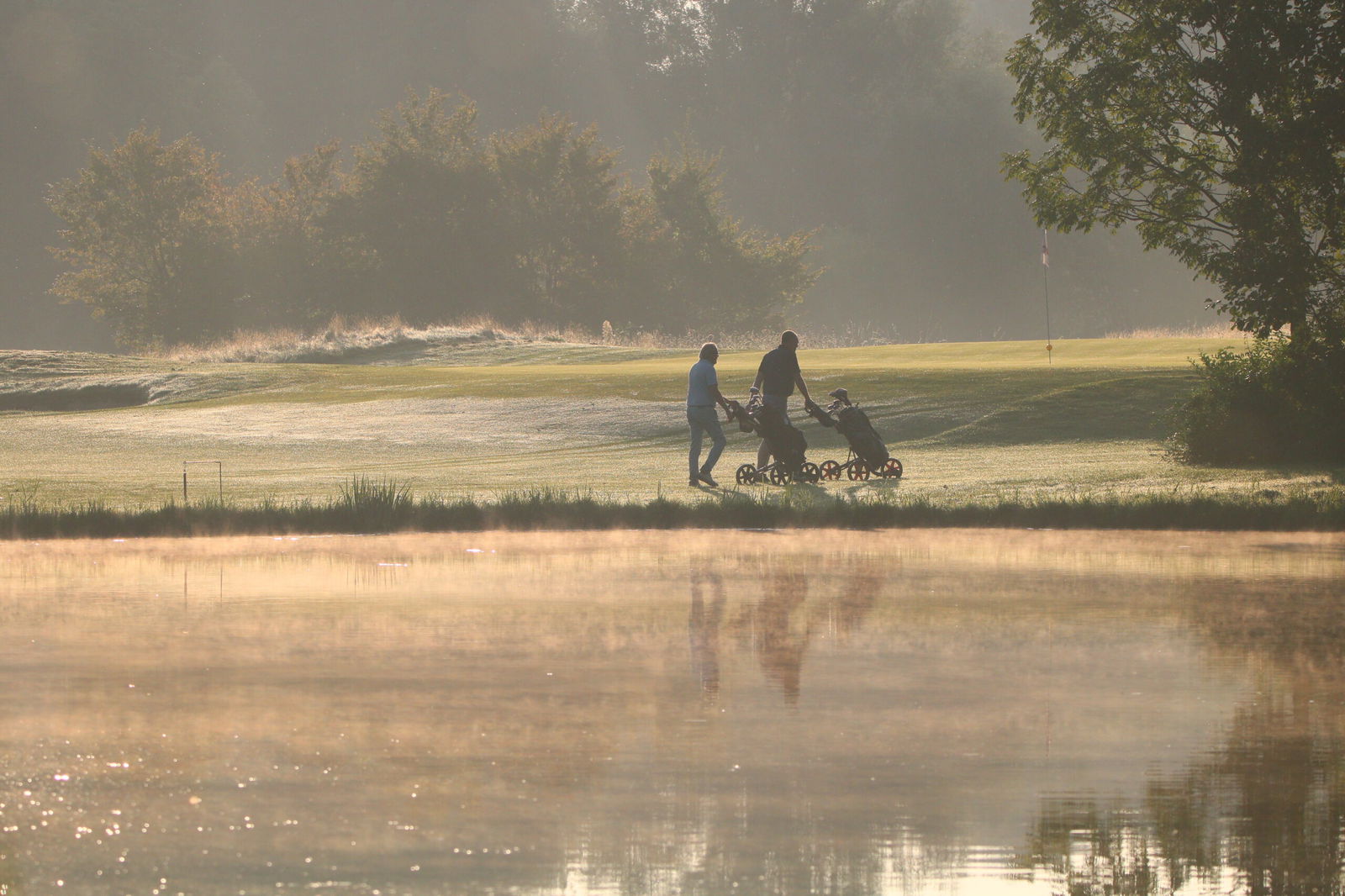 Golfers lopen met trolleys langs de golfbaan bij zonsopkomst met water en mist